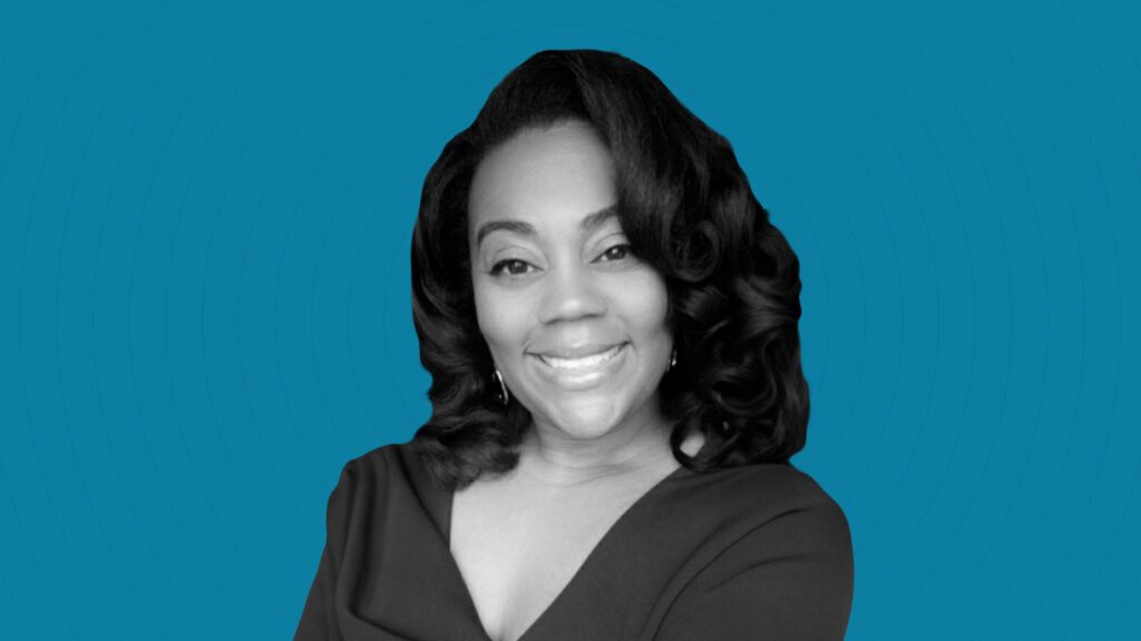 A black and white image of Kamilah Lewis smiling against a deep blue background.