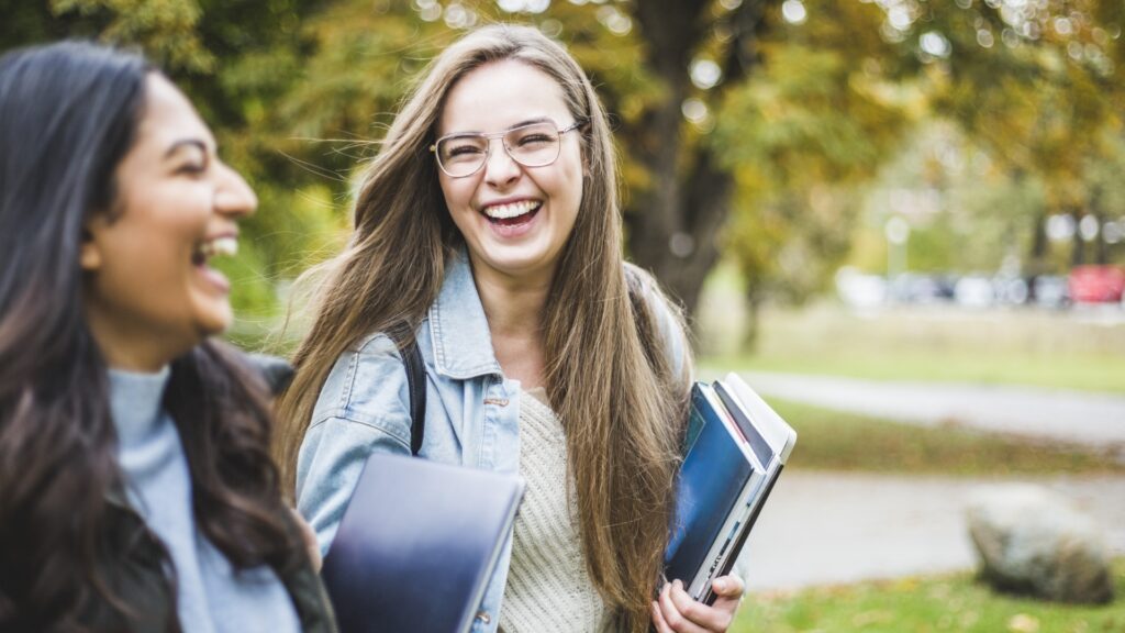 Two students walk across campus carrying books and laughing together.