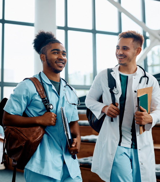 Two medical students walk through a building wearing backpacks and stethoscopes while holding notebooks.