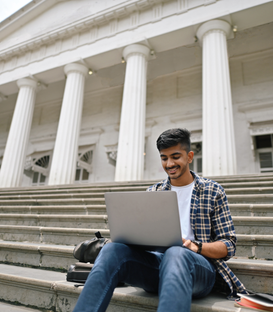 A man sits outside on the steps of a columned building while working on a laptop.