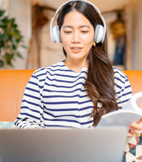 A woman wearing headphones holds a book and sits in front of a laptop screen.