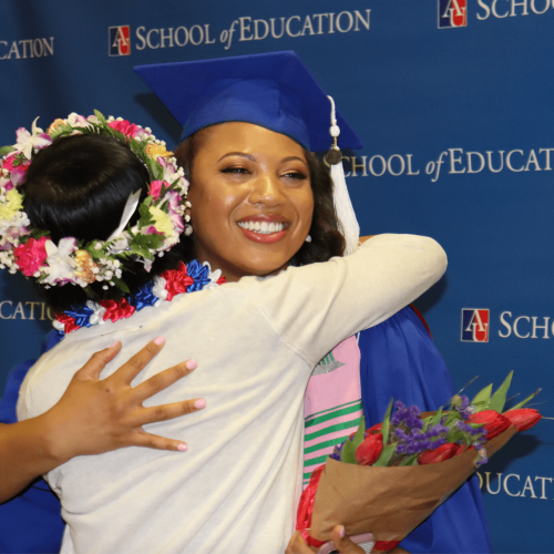 A graduate wearing a cap and gown hugs another person in front of a step-and-repeat.