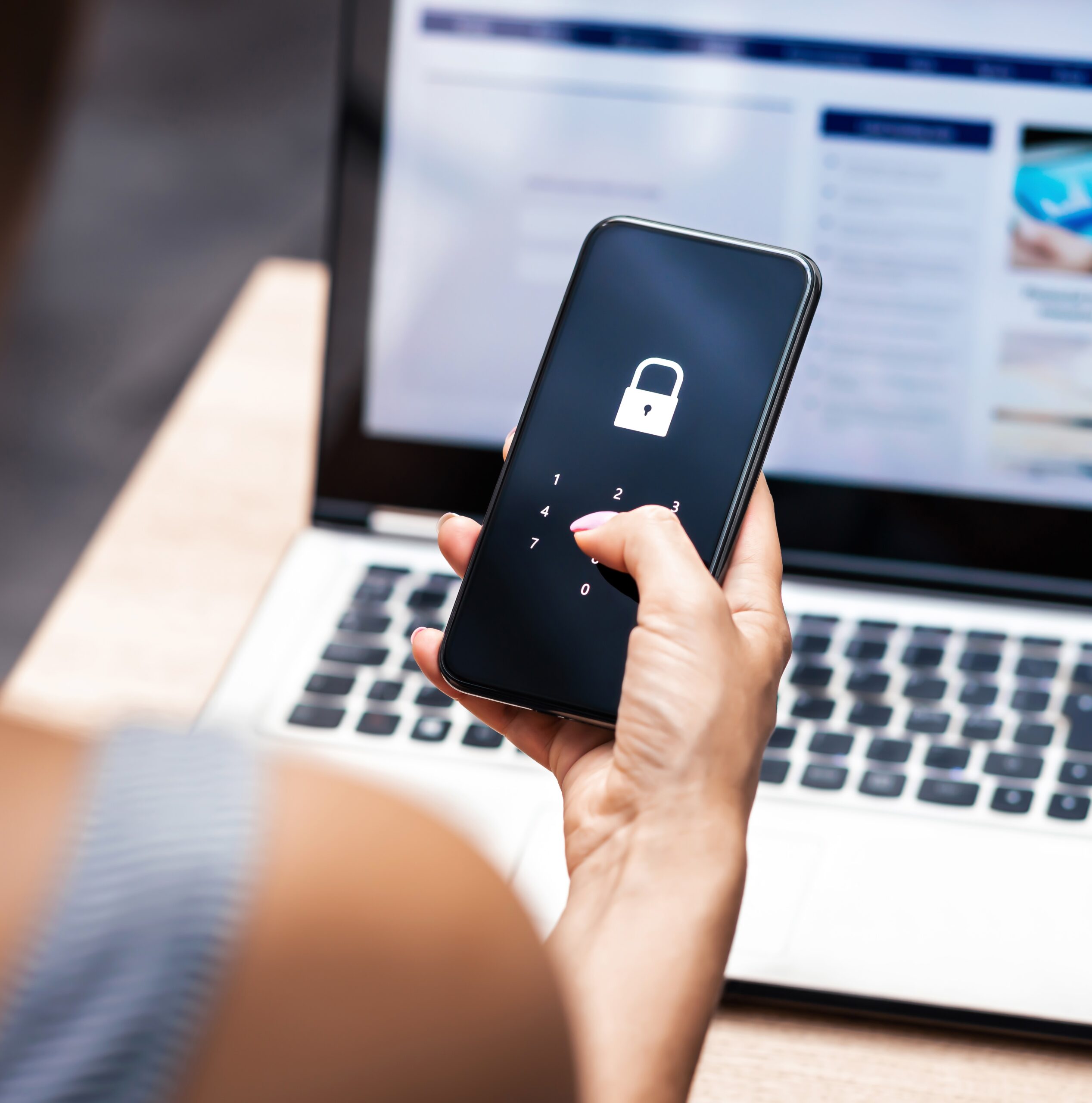 A woman holds a smart phone with a lock screen in front of an open laptop.