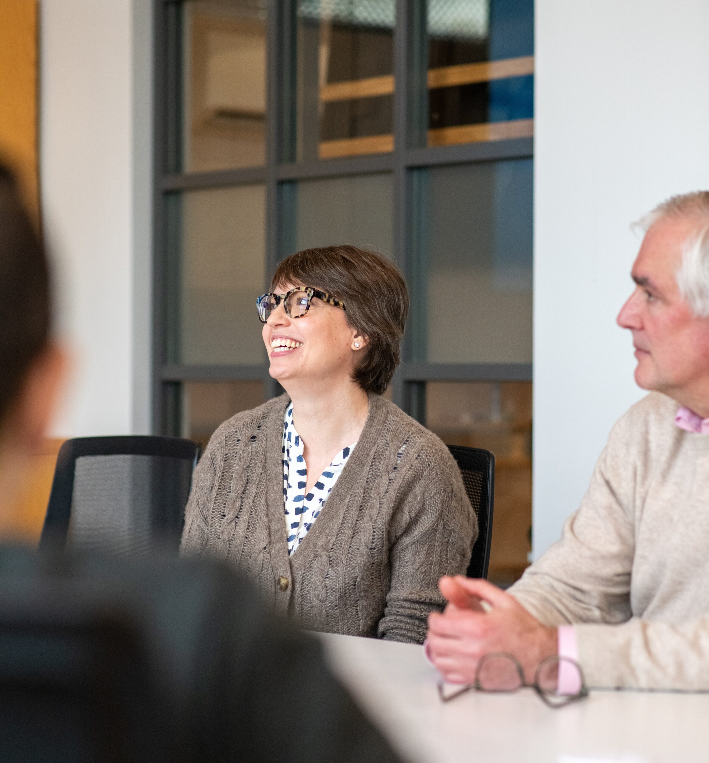 A woman wearing glasses smiles as she sits beside a colleague at a conference table in an open concept office.