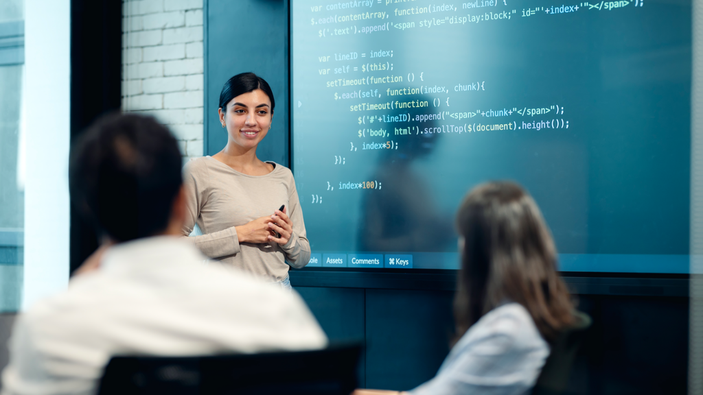 A student presents to her class in front of a large screen containing lines of code.