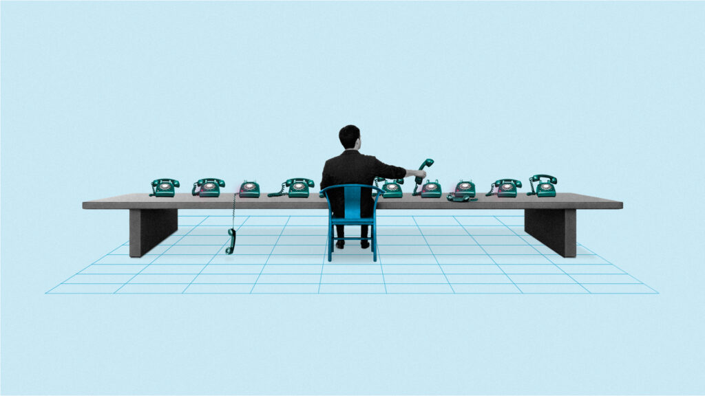 A man sits at a long table answering multiple rotary phones, each placed in a row.